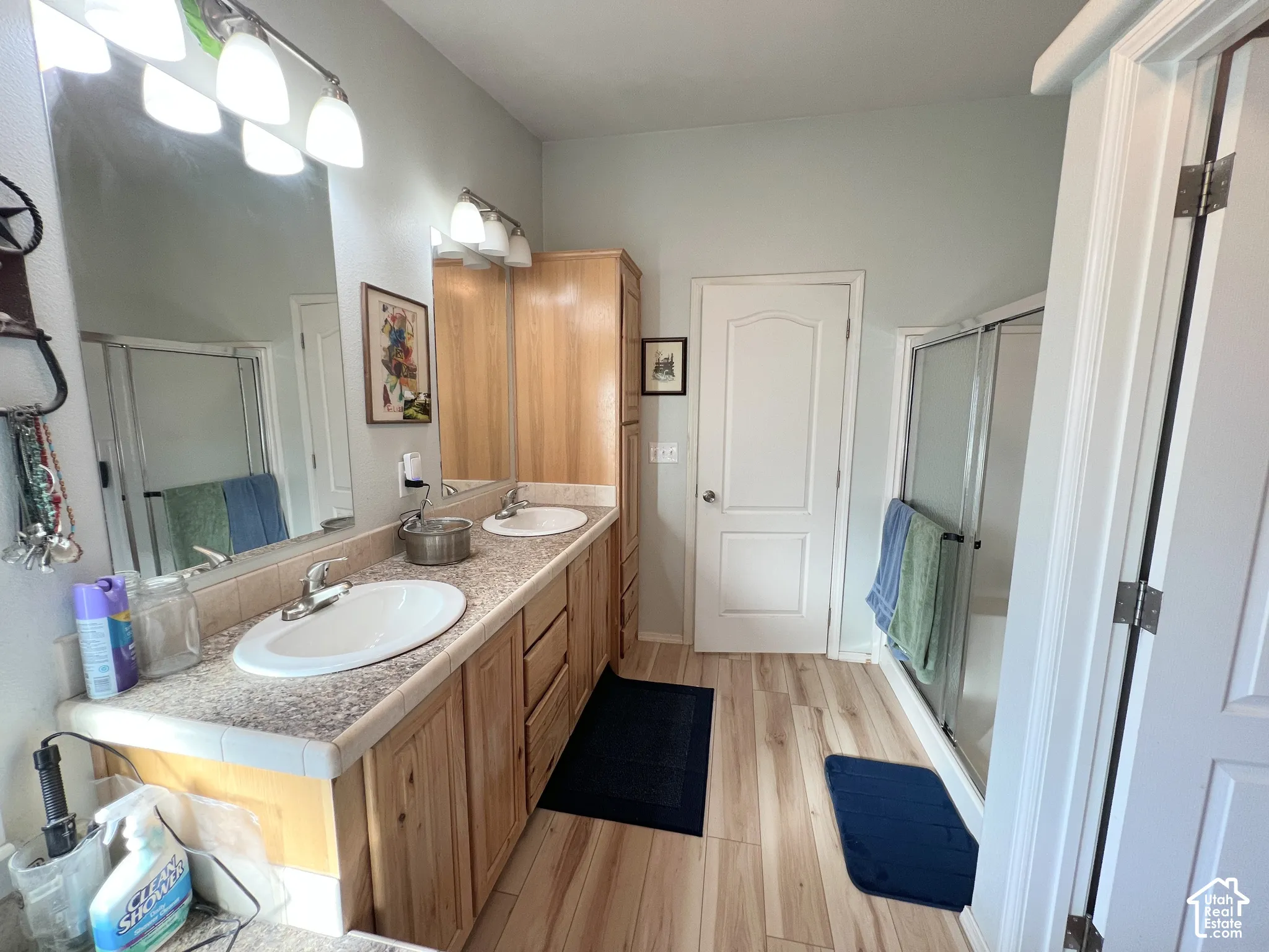 Dual vanity with laminate countertops, wood-finish flooring, built-in wood cabinetry, and a shower with a sliding glass enclosure