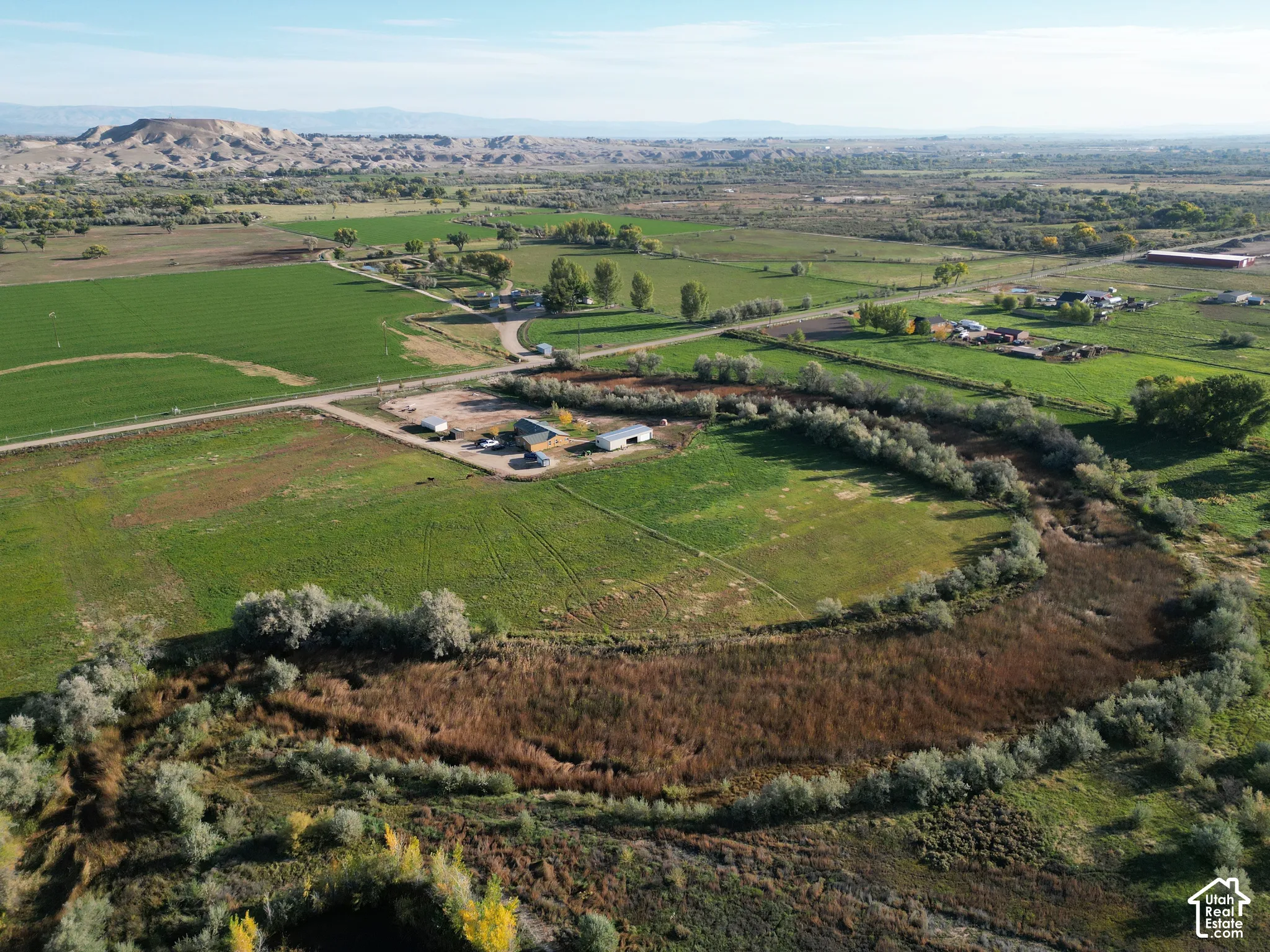 Expansive agricultural land featuring a meandering watercourse, multiple structures, established trees, and distant natural terrain