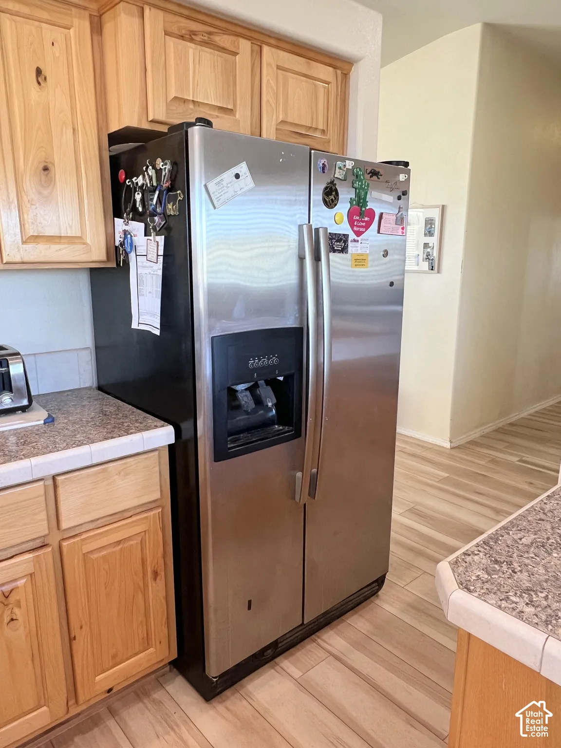 Stainless steel side-by-side refrigerator, light wood-finish cabinetry, speckled countertop with tile edge, and wood-finish flooring