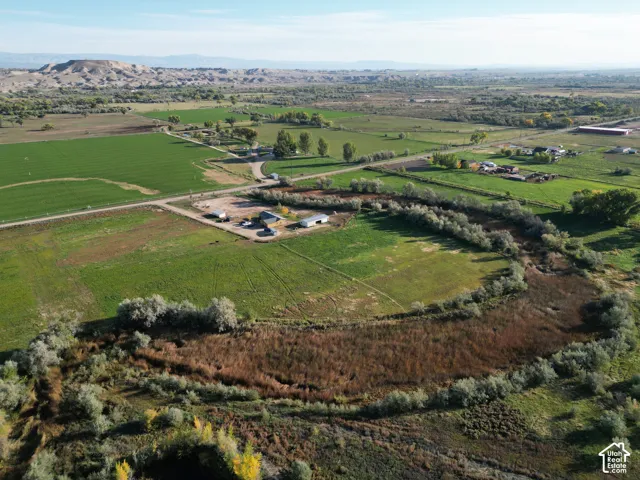 Expansive agricultural land featuring a meandering watercourse, multiple structures, established trees, and distant natural terrain
