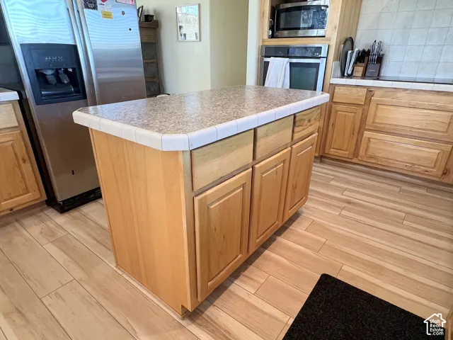 Kitchen featuring an island with tile-edged countertop, wood-finish flooring, built-in stainless steel appliances, and natural wood cabinetry