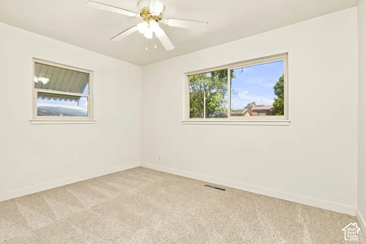 Soft neutral carpet flooring, a ceiling fan with integrated lighting, and two windows providing natural illumination