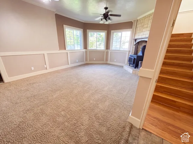 Carpeted living area with wainscoting, multiple windows, a ceiling fan, and a fireplace with a wood-burning stove