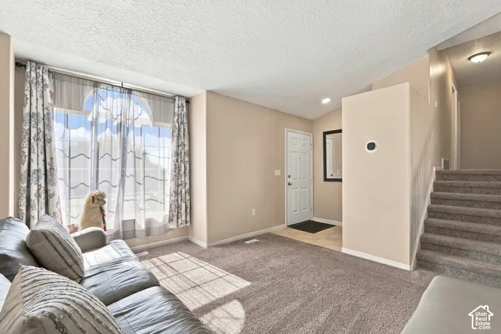 Bright living area with expansive windows featuring sheer drapery and natural light, neutral wall finishes, carpeted flooring, tile entryway, and direct access to a staircase