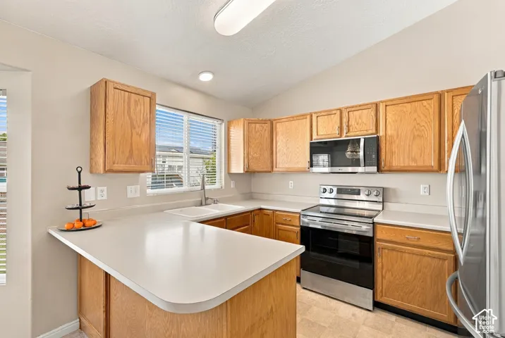 Kitchen featuring natural wood cabinetry, light-toned countertops, stainless steel appliances, a double basin sink, and vaulted ceilings