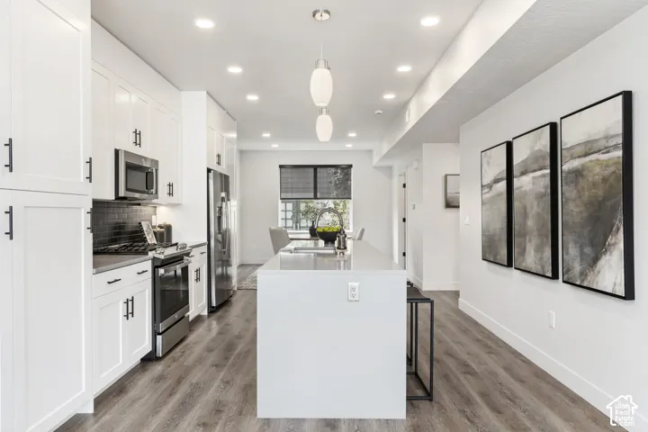 Open-concept kitchen featuring white cabinetry, stainless steel appliances, a dark subway tile backsplash, a large center island, and wood-finish flooring Pictures are of the model home