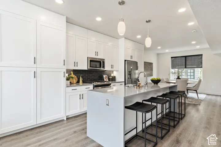 Kitchen featuring white shaker cabinetry, stainless steel appliances, a dark subway tile backsplash, a large center island with a sink, and wood-finish flooring Pictures are of the model home