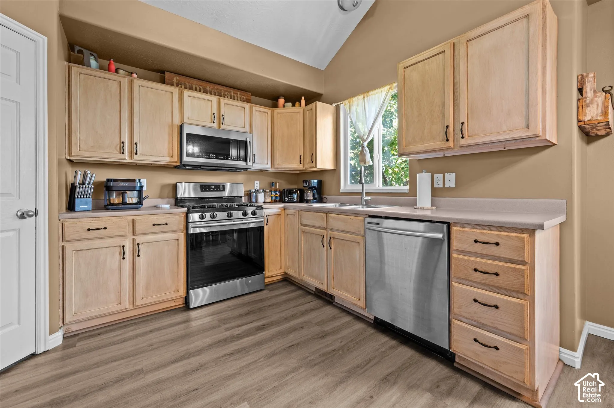 Kitchen featuring wood-finish flooring, light wood cabinetry, stainless steel appliances, and a vaulted ceiling