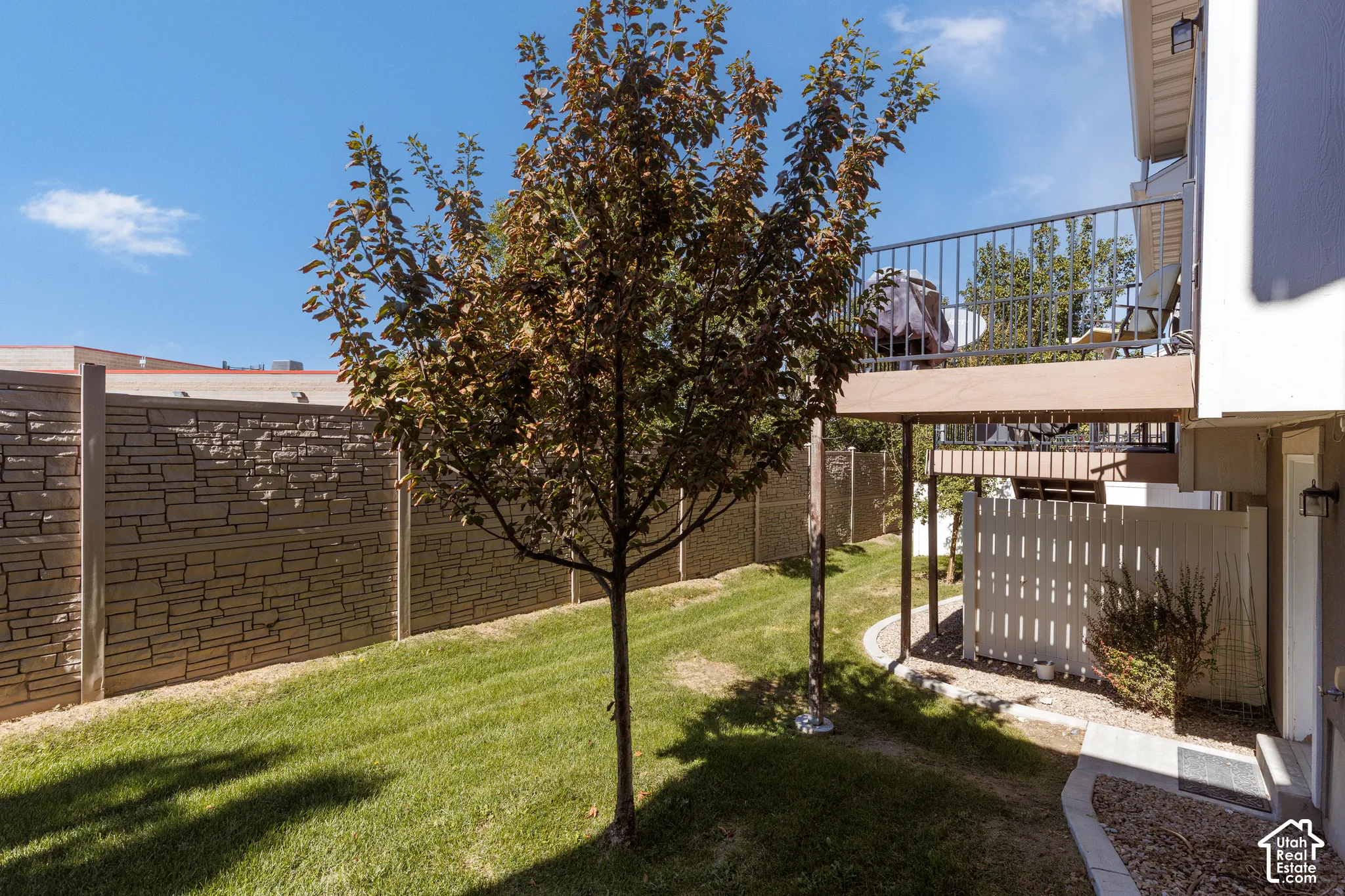 Manicured lawn, mature tree, stone-patterned privacy fence, elevated balcony with metal railing, and exterior wall sconces