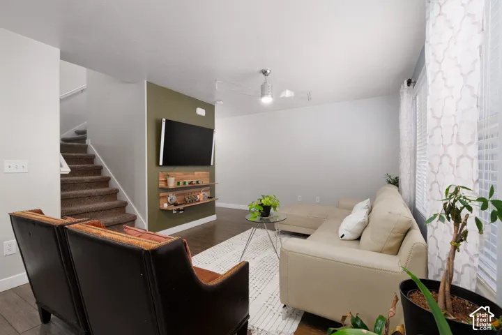 Wood-finish flooring, staircase with carpeted treads, wall-mounted shelving, multiple windows with blinds, and a ceiling fan with integrated lighting