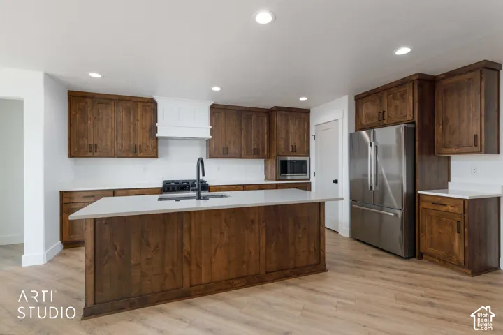 Kitchen featuring dark wood cabinetry, light two-tone countertops, stainless steel appliances, a center island with a sink, and wood-finish flooring