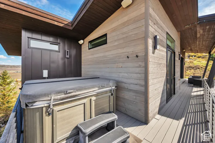 Modern deck featuring composite decking, integrated hot tub with built-in seating and insulated cover, horizontal wood-finish siding, black framed glass doors, and expansive open-air layout with scenic backdrop