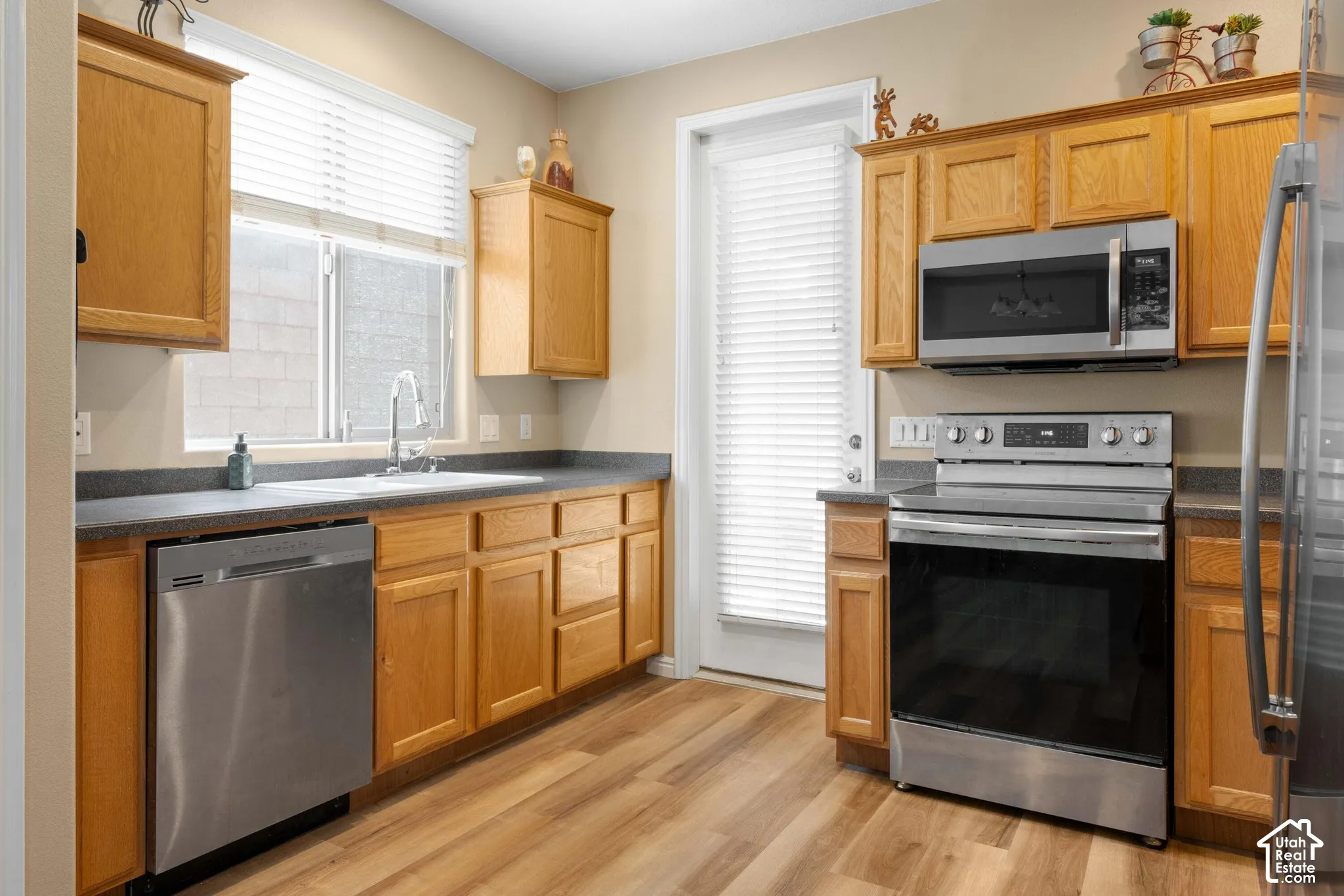 Kitchen featuring wood-finish flooring, oak cabinetry, stainless steel appliances, a double basin sink, and a window with blinds