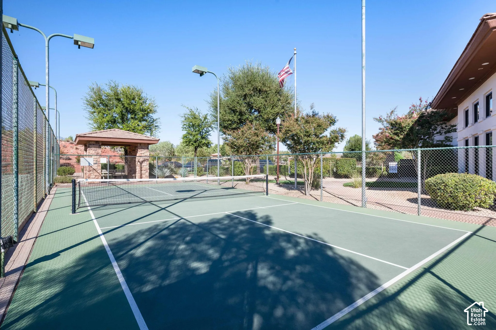 Outdoor tennis court with chain-link fencing, court lighting, and a stone-clad shaded structure