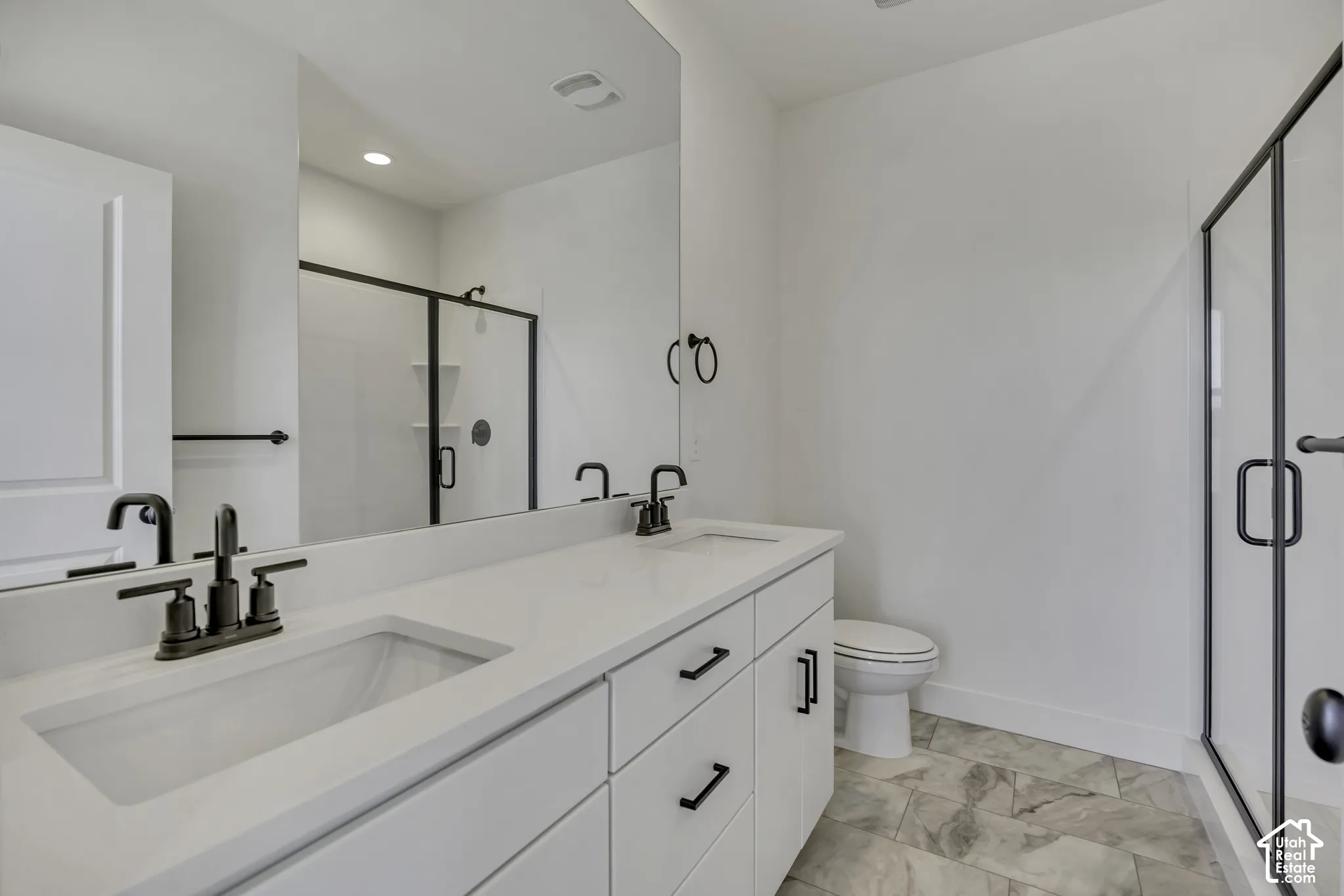Double vanity with integrated sinks, white cabinetry, matte black hardware, glass-enclosed shower, and large format tile flooring