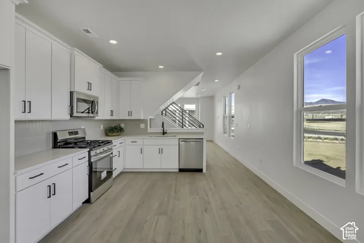 Kitchen with white cabinetry, stainless steel appliances, light-toned wood-finish flooring, recessed lighting, and a staircase with metal railings