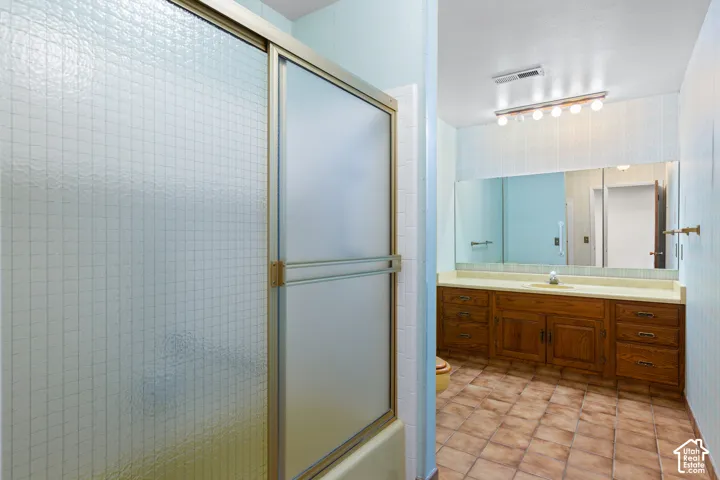 Full-length vanity mirror, wood-finish cabinetry, light-toned countertop, ceramic tile flooring, and frosted glass shower enclosure
