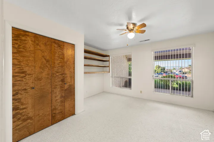 Carpeted flooring, wood-finish bi-fold closet doors, built-in shelving, ceiling fan, and two windows with vertical blinds