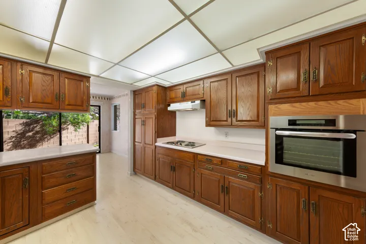 Wood cabinetry with brass hardware, built-in stainless steel oven, white countertops, built-in cooktop, and light wood-finish flooring