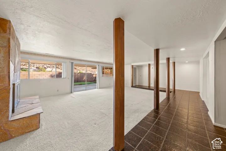 Spacious room featuring a stone-clad fireplace, extensive window line, sliding glass door, exposed support beams, and a combination of carpeting and tile flooring