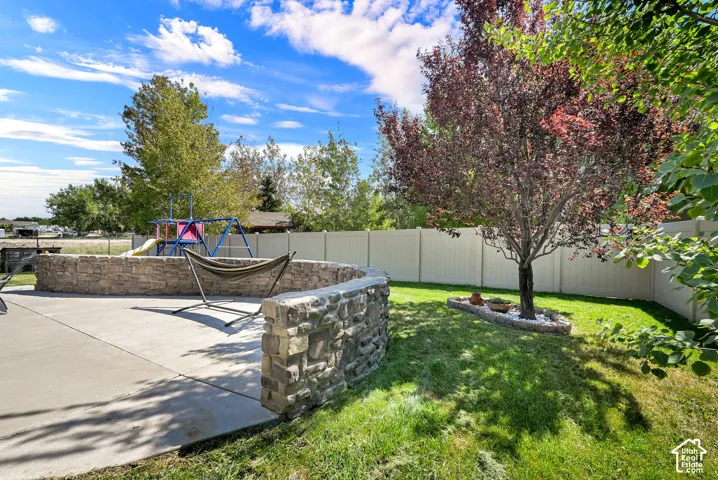 Expansive concrete patio bordered by a stone retaining wall, featuring a manicured lawn, established shade trees, and a privacy fence