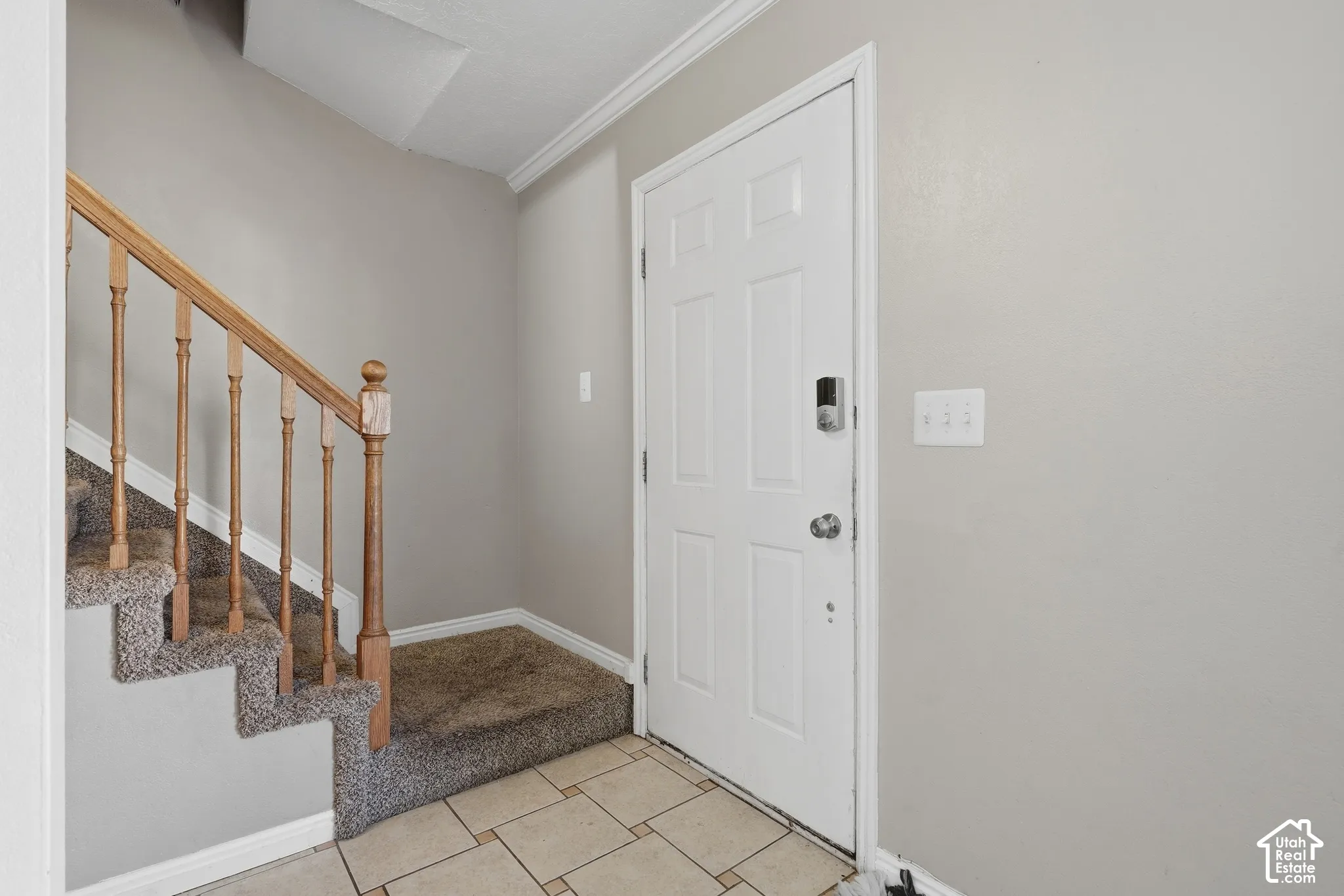 Entryway featuring tiled flooring, carpeted staircase with wood railing, crown molding, and a paneled door with smart lock