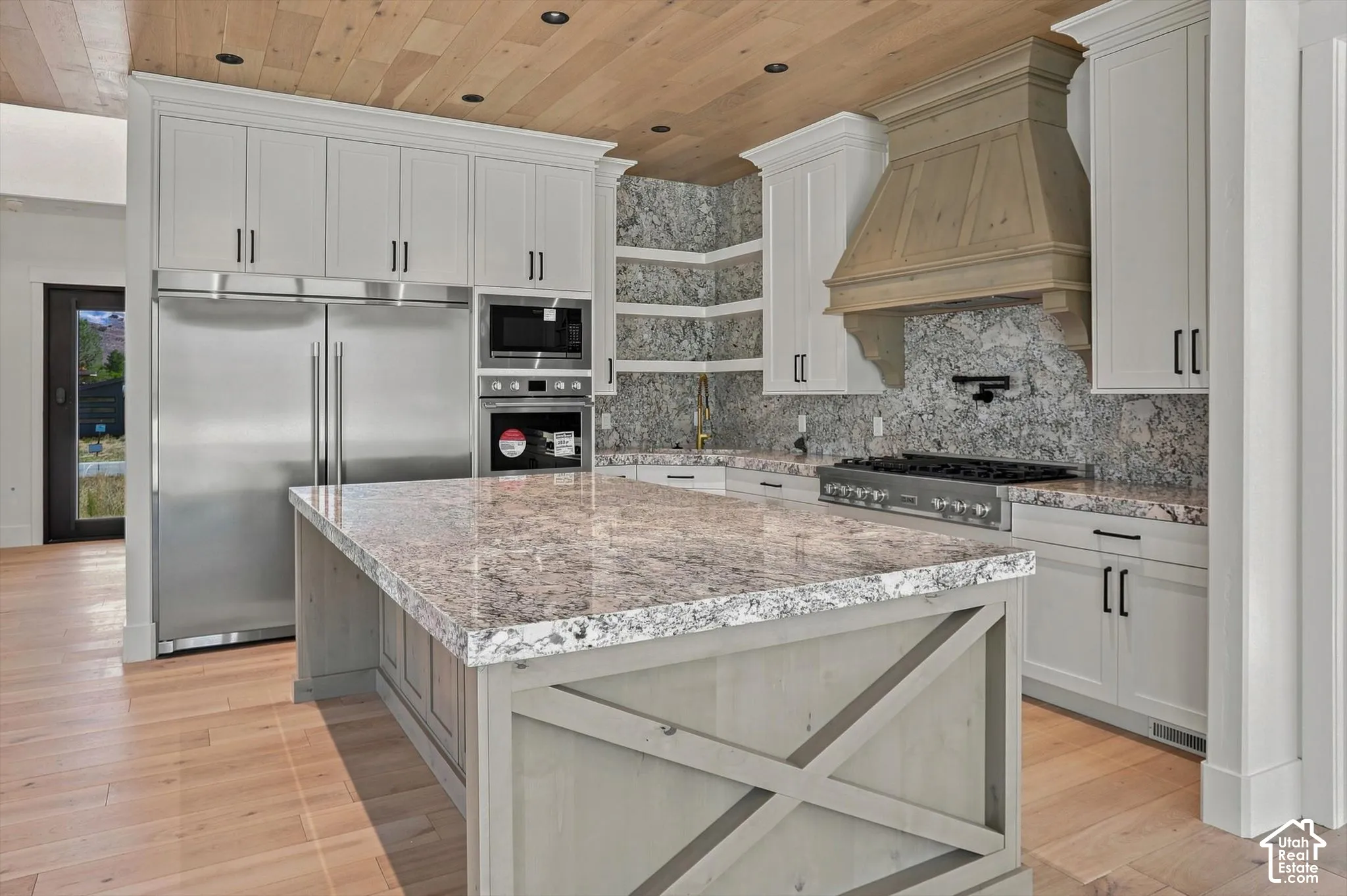 Gourmet kitchen featuring a large island with a stone countertop, light wood-finish flooring, white cabinetry with dark hardware, stainless steel appliances, and a wood-paneled ceiling