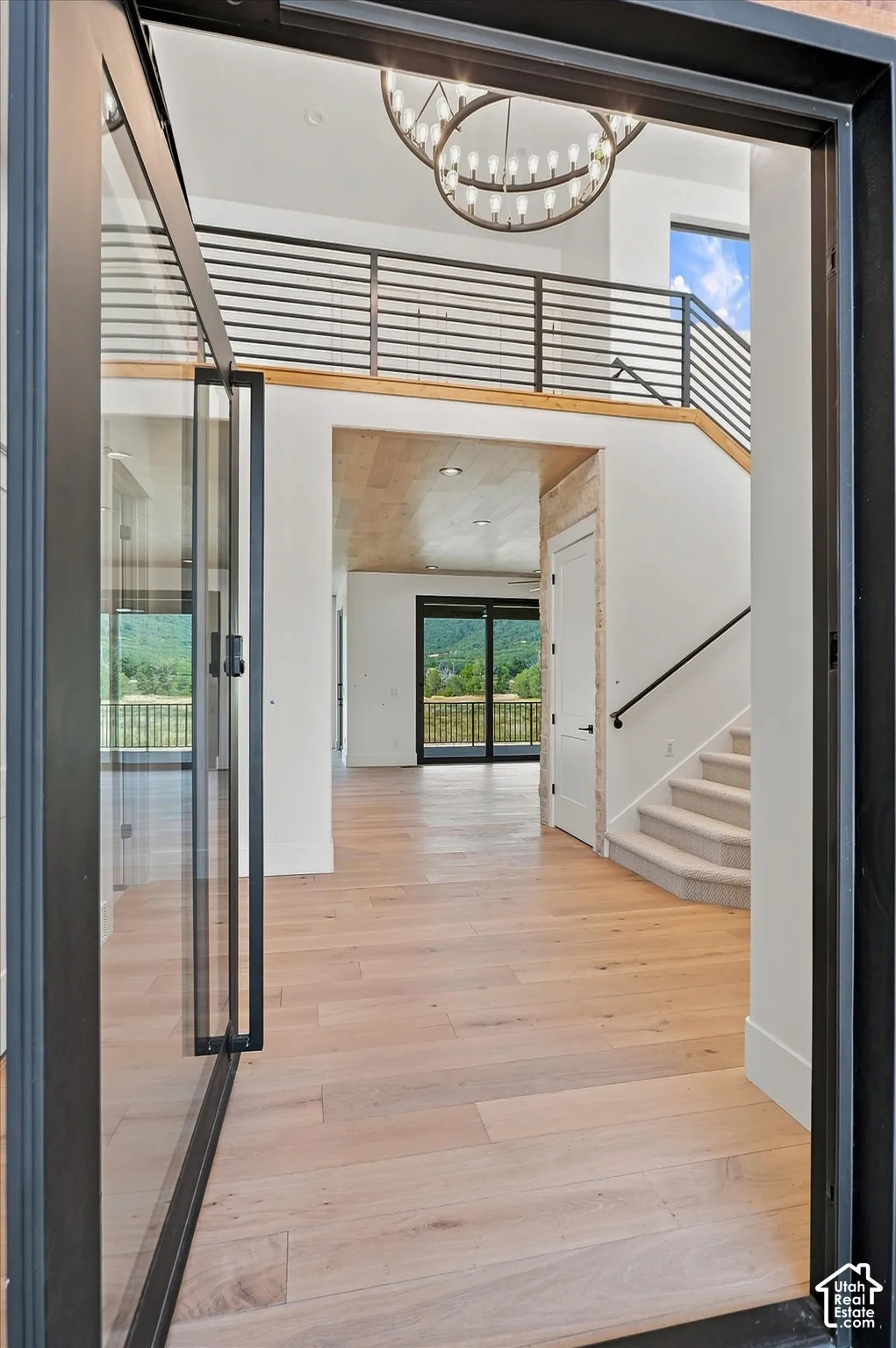 Light wood-finish flooring, a two-story entryway, a circular chandelier, open-tread staircase with metal railing, and a glass pivot door