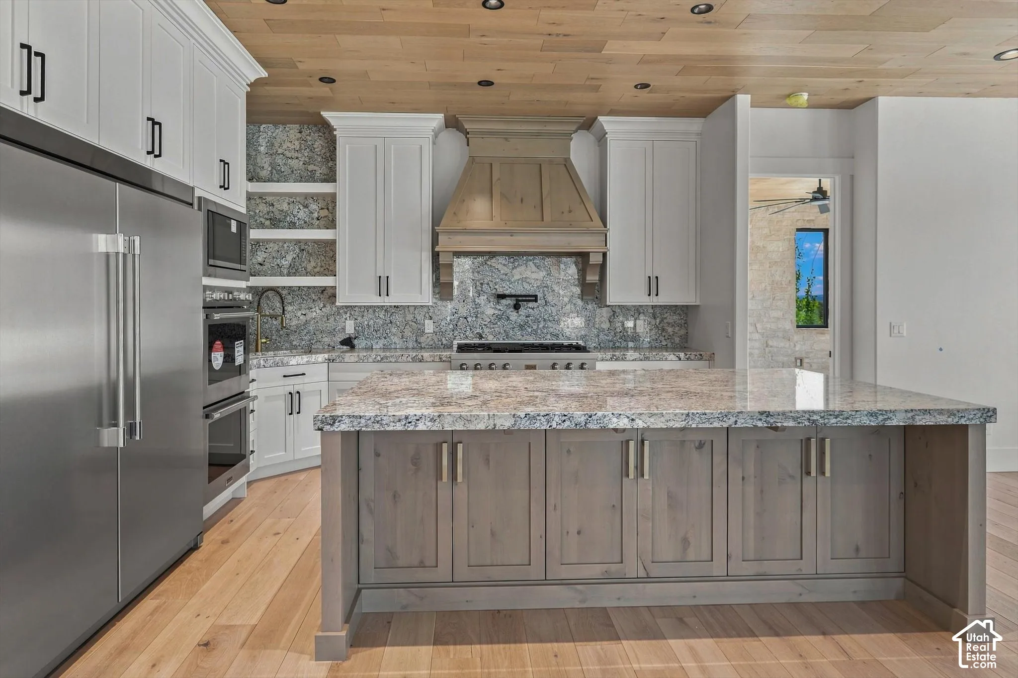 Gourmet kitchen with stainless steel appliances, light wood-finish flooring, white upper cabinetry, a central island with a light-toned countertop, and a wood-paneled ceiling