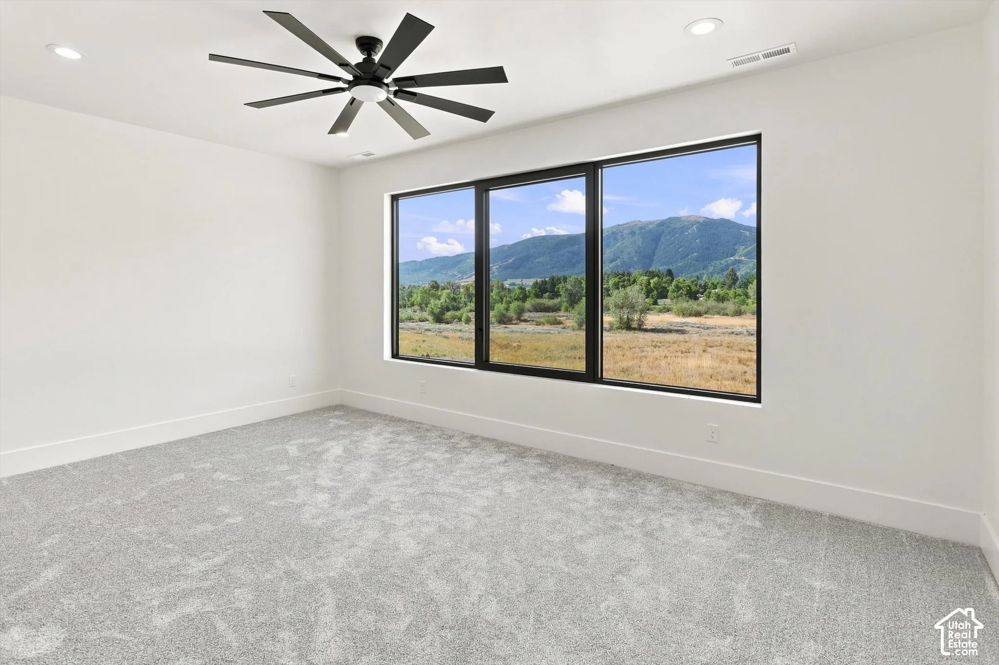 Expansive windows featuring black frames, recessed ceiling lights, a multi-blade ceiling fan, and light grey carpeting