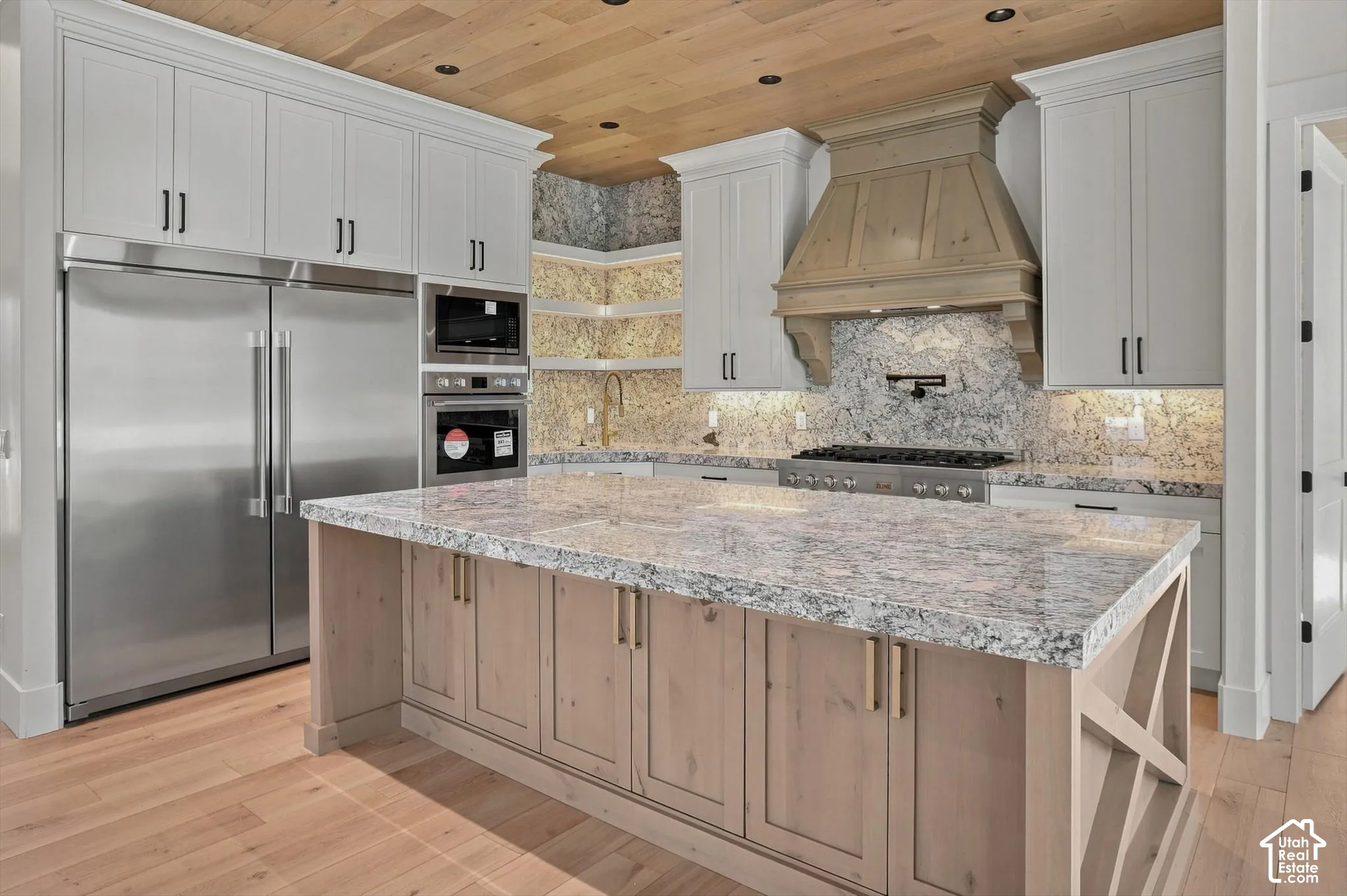 Gourmet kitchen featuring a large central island with a stone countertop, light wood-finish cabinetry, stainless steel appliances, and a wood-paneled ceiling