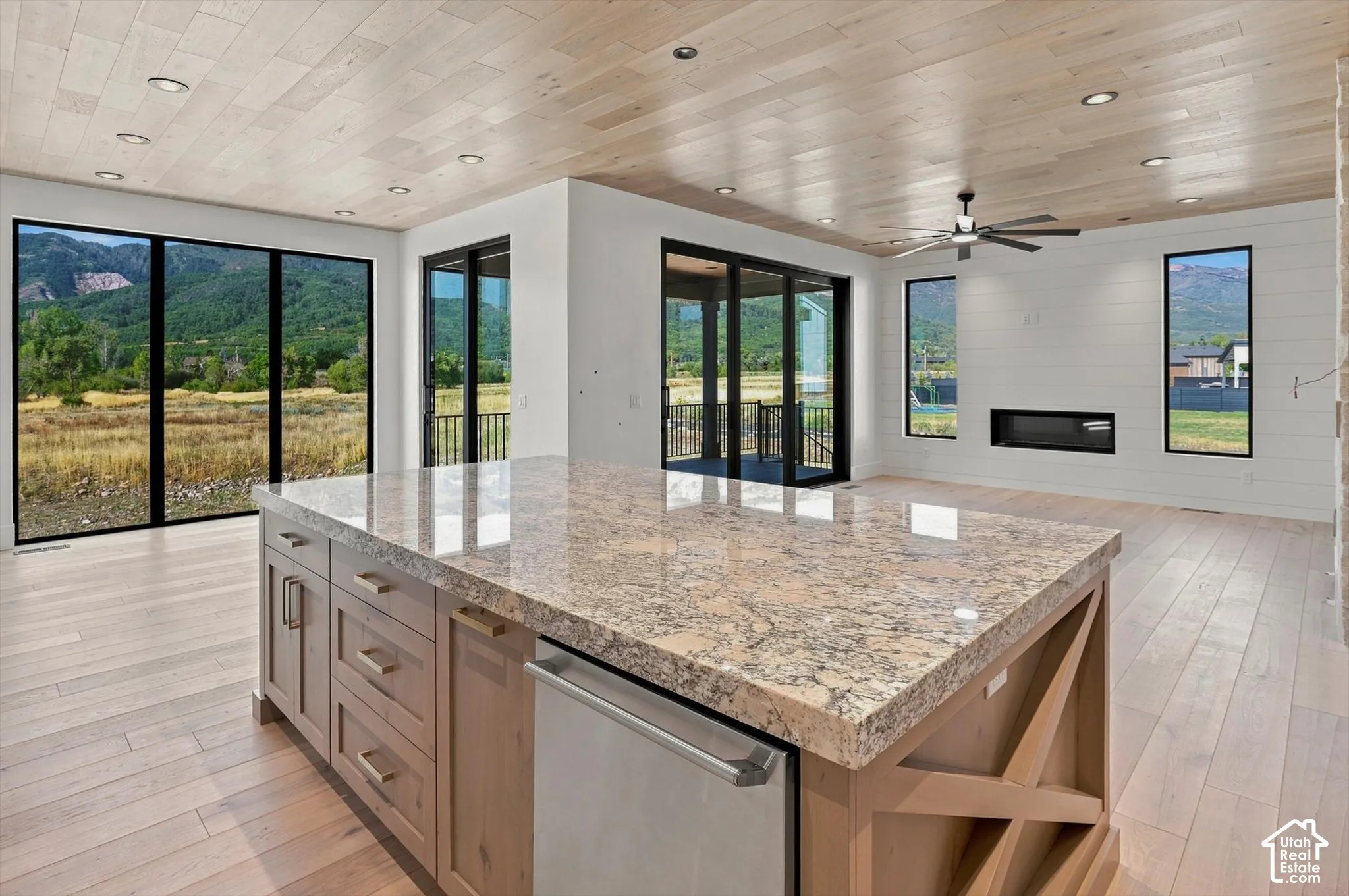 Spacious open-concept interior with light wood-finish flooring and ceiling, a substantial kitchen island with a stone countertop, an integrated modern fireplace, and large windows