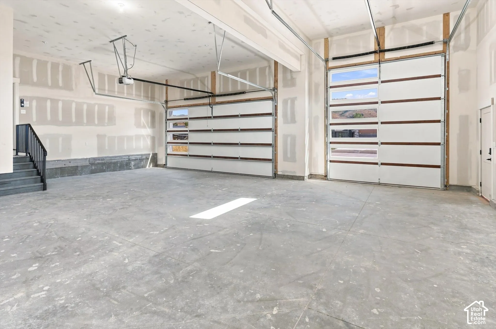 Two garage doors with horizontal window inserts, concrete flooring, and a staircase with a black metal handrail