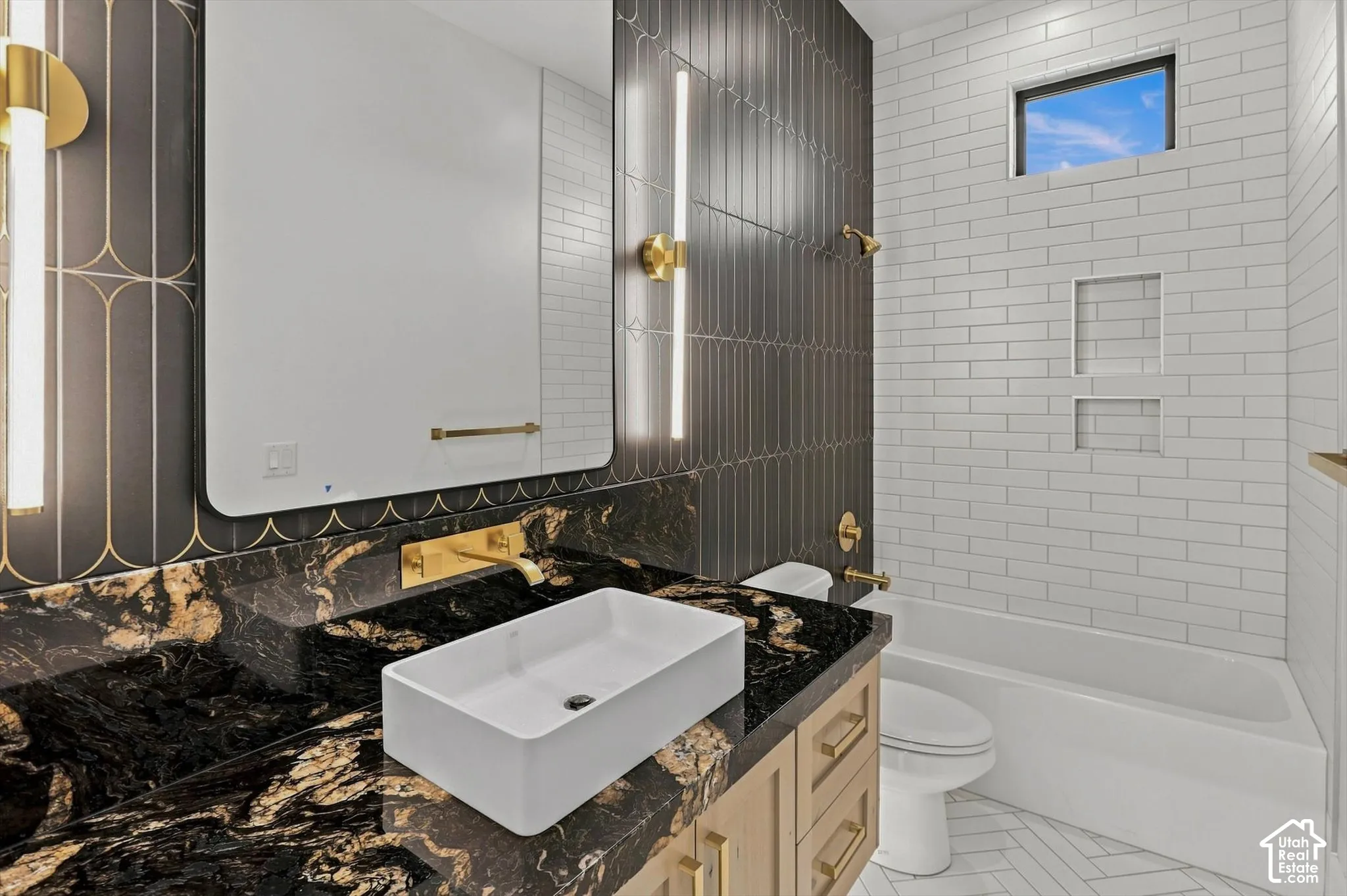 Black and gold patterned wall tile, rectangular vessel sink, wall-mounted gold faucet, black stone vanity top, and light wood-finish cabinetry with gold hardware