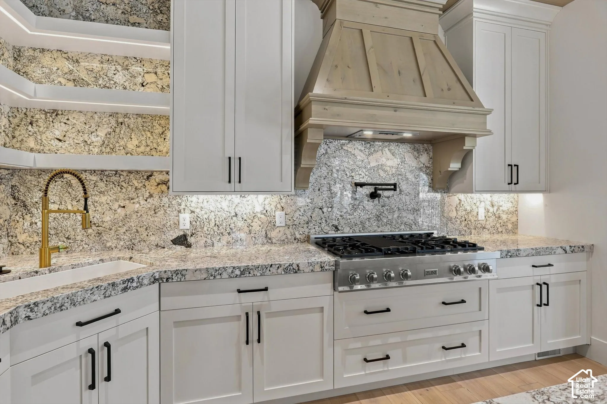 White shaker-style cabinetry with matte black hardware, granite countertops and full backsplash, built-in stainless steel range, and a brass-finish gooseneck faucet