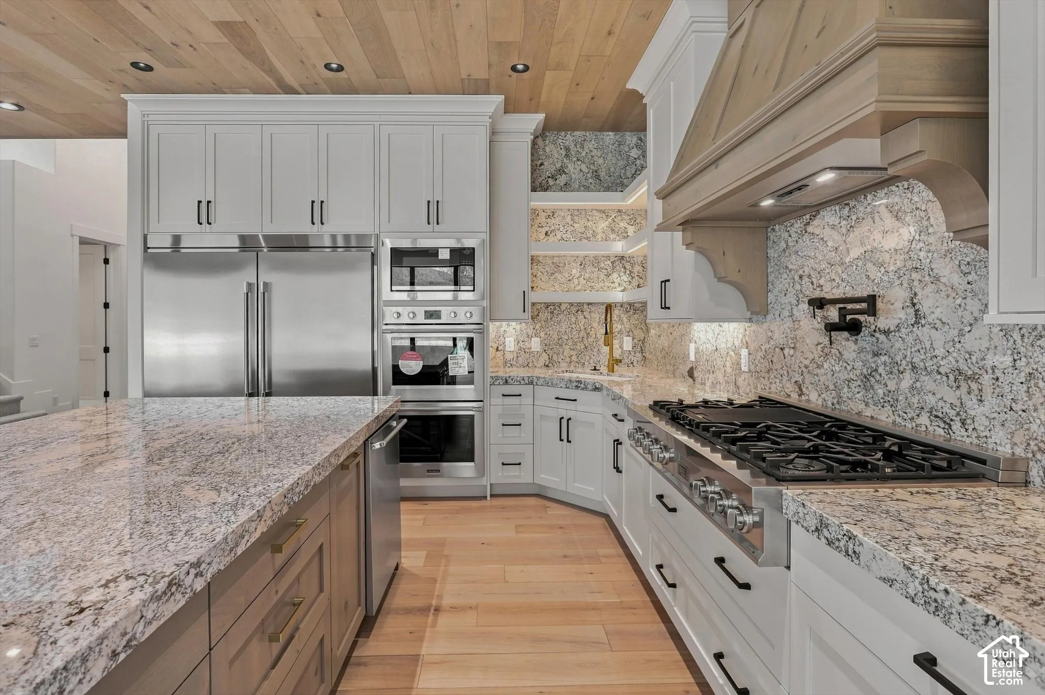Gourmet kitchen with natural wood plank ceiling, light-toned cabinetry, integrated stainless steel appliances, a pot filler, and speckled stone countertops