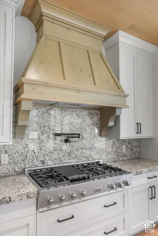 Wood-finish range hood, stainless steel range, mottled stone backsplash, light-toned countertops, and white cabinetry with dark hardware