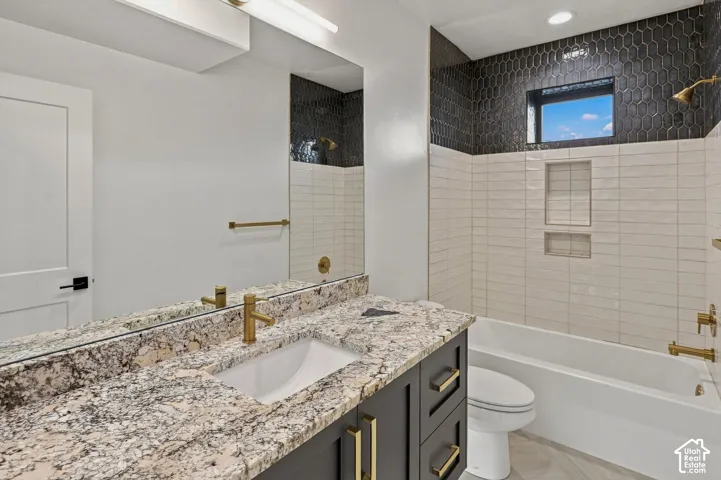 Vanity with stone countertop and integrated sink, gold-finish fixtures, dark cabinetry with gold hardware, full-width mirror, and a tiled bathtub with built-in niches