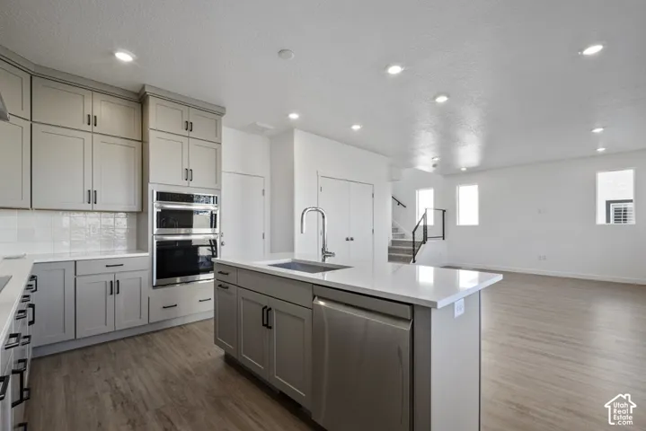 Kitchen featuring gray cabinetry, stainless steel double wall ovens, a center island with a sink, light countertops, and wood-finish flooring