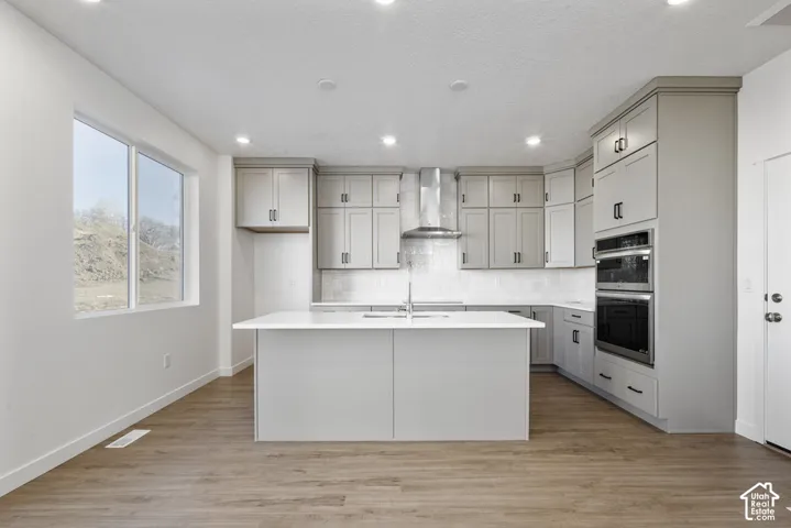 Modern kitchen featuring gray cabinetry, stainless steel appliances, a white tile backsplash, a central island with a sink, and wood-finish flooring