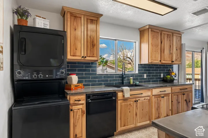 Wood-finish cabinetry, subway tile backsplash, dark countertops, black appliances, and a built-in laundry stack