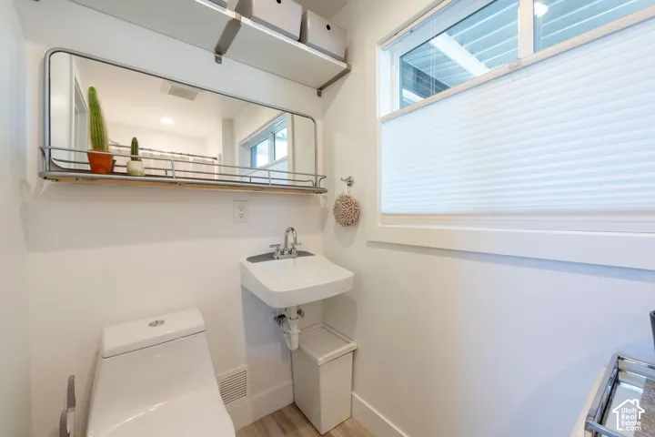 Wall-mounted mirror with integrated shelf, open shelving, white wall-mounted sink, white toilet, and window with privacy blinds