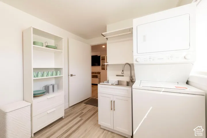 White stacked laundry appliances, stainless steel utility sink with chrome faucet, white cabinetry, wood-finish flooring, and a white flat-panel door