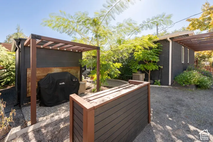 Gravel hardscaping, planter box with composite wood-finish, pergola with a wood-finish frame, exterior building with black siding, and mature landscaping