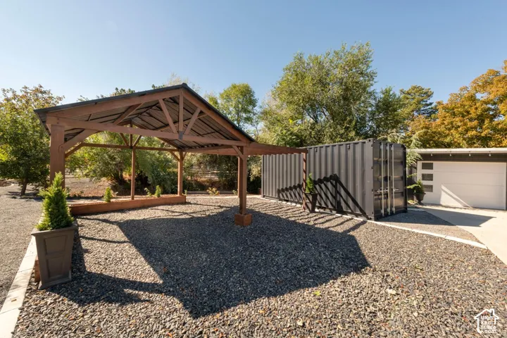 Gravel driveway with a wood pergola, dark grey corrugated metal structure, white garage door with horizontal windows, and mature trees