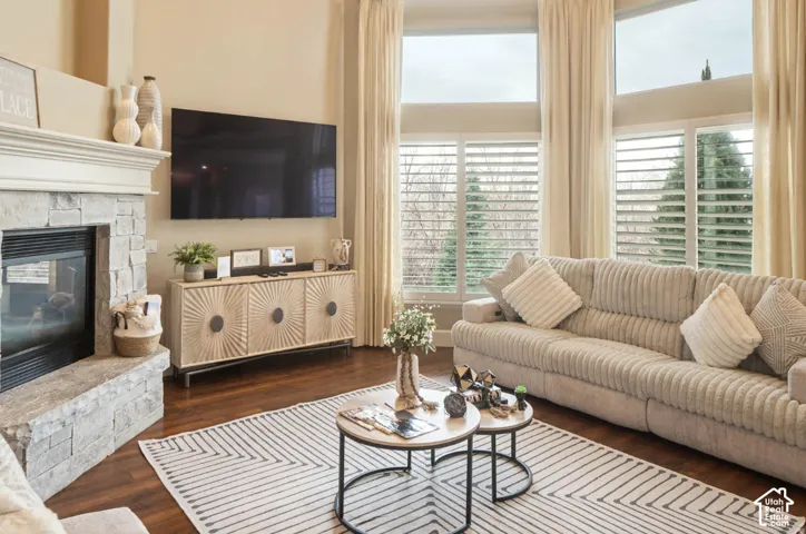 Stone-clad fireplace with mantel, dark wood-finish flooring, high ceilings, tall windows with plantation shutters, and a media console