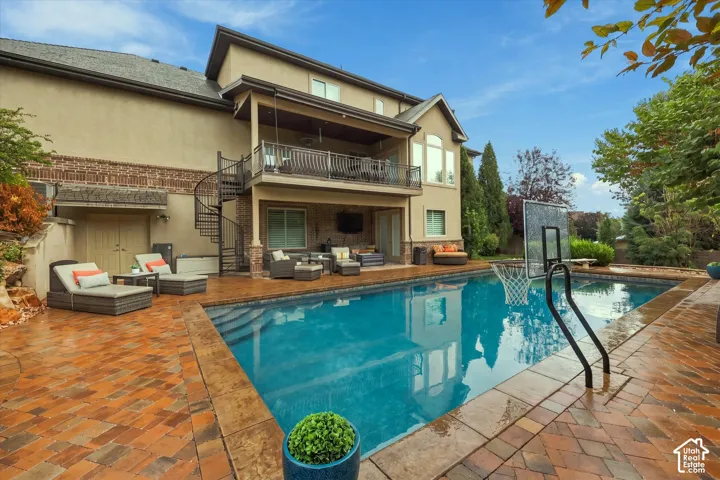 Custom pool with integrated basketball hoop, extensive paver patio, covered balcony, spiral staircase, and stucco exterior