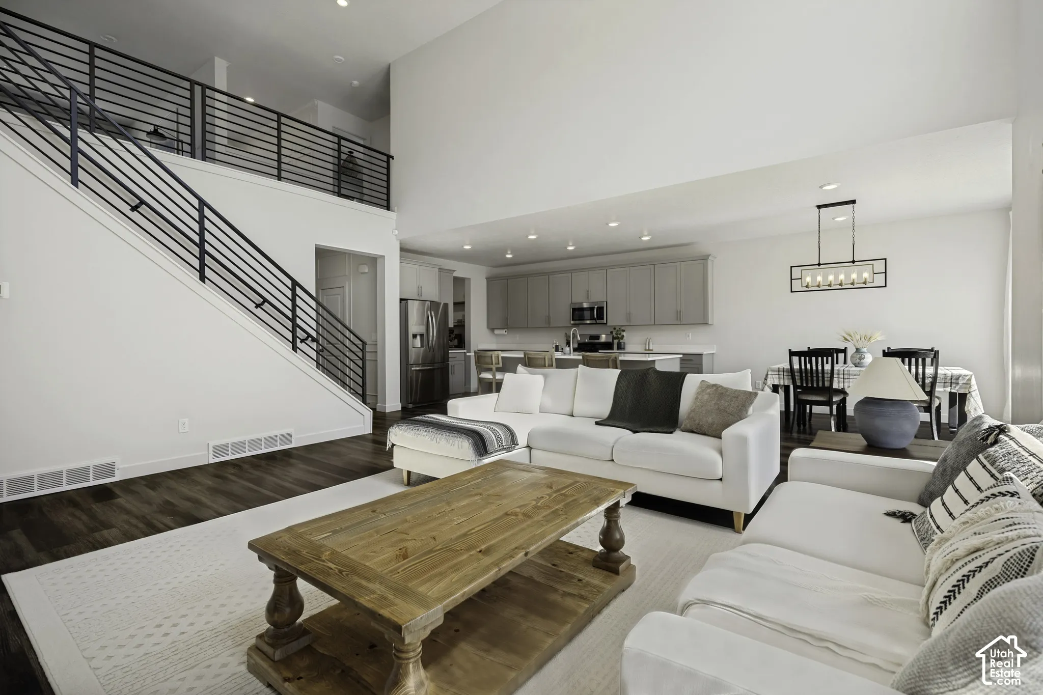 Open-concept living area with soaring ceilings, wood-finish flooring, a modern staircase with black horizontal railings, an integrated kitchen with stainless steel appliances, and a dining area with a rectangular chandelier