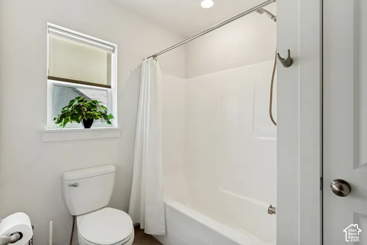 Bathroom featuring a white toilet, window with roller shade, and a shower/tub combination with a wall-mounted showerhead