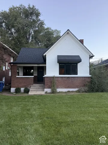 White stucco and brick exterior, dark asphalt shingle roof, front porch with steps, and a dark awning over a window